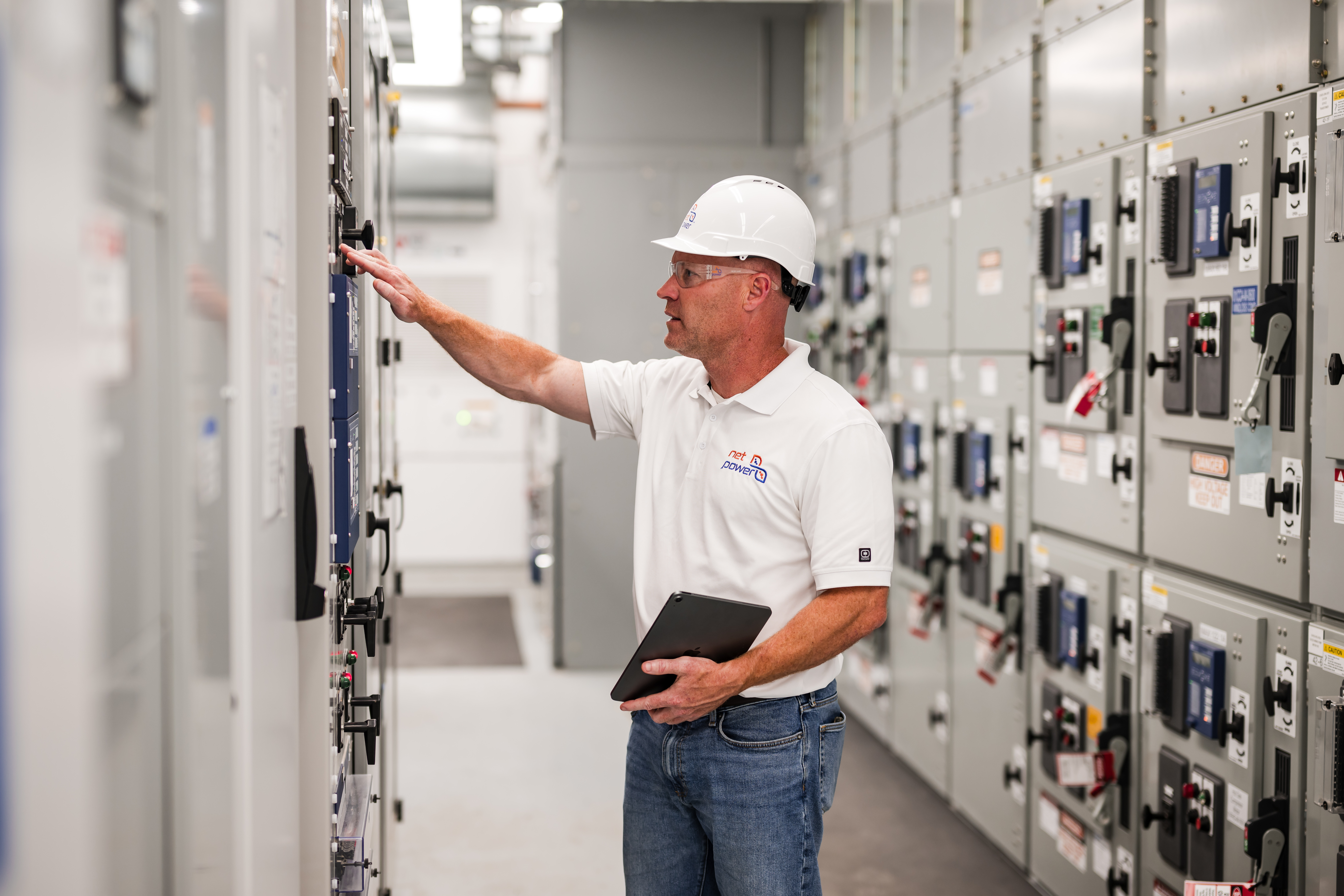Man in a white hard hat and polo shirt interacting with electrical panels in a well-lit industrial room.